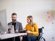 Two people sitting at a desk looking at a computer. One of them is a wheelchair user.