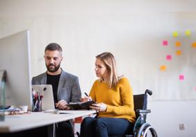 Two people sitting at a desk looking at a computer. One of them is a wheelchair user.