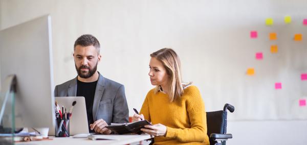 Two people sitting at a desk looking at a computer. One of them is a wheelchair user.