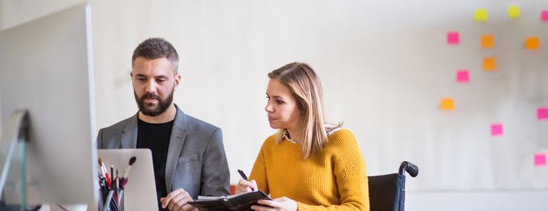 Two people sitting at a desk looking at a computer. One of them is a wheelchair user.