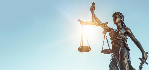 A bronze statue of Lady Justice holding balanced scales in her left hand and a sword in her right, with sunlight shining through the scales against a clear blue sky.