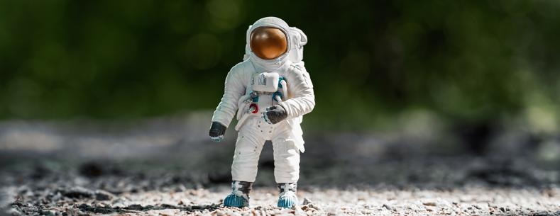 The image shows a white toy astronaut on dirt with trees in the background.