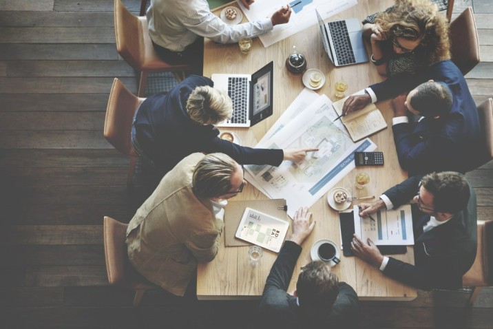 An image from above with smartly dressed people sat around a table with laptops and paper plans discussing and pointing at the plans.