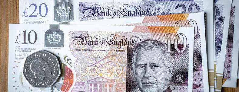 Ten pound sterling note on top of other notes, a 50 pence piece is on top of the ten pound note. The note and coin carry the image of the King.