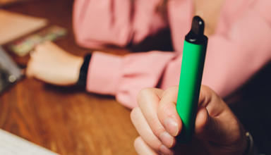 A person holding a green vape pen or e-cigarette. The focus is on the hand and the vape device 