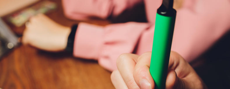 A person holding a green vape pen or e-cigarette. The focus is on the hand and the vape device 