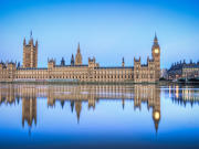 The UK Houses of Parliament in daytime from across the River Thames, with its reflection showing in the water. 