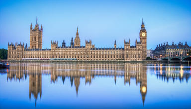 The UK Houses of Parliament in daytime from across the River Thames, with its reflection showing in the water. 