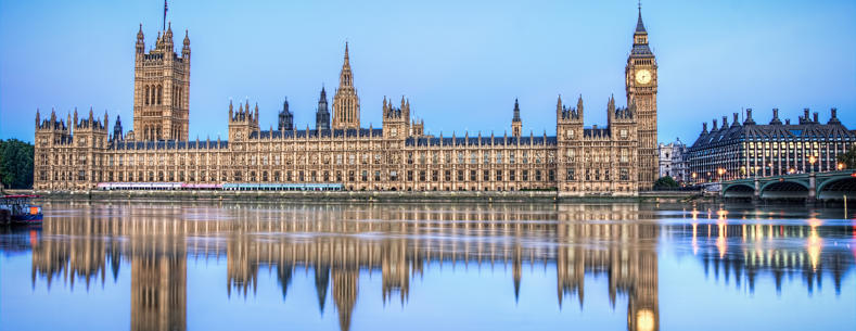 The UK Houses of Parliament in daytime from across the River Thames, with its reflection showing in the water.
