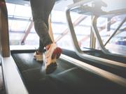 A person’s legs wearing sports trousers and trainers running on a treadmill with windows in the background.