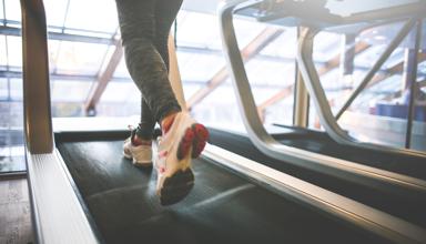 A person’s legs wearing sports trousers and trainers running on a treadmill with windows in the background.