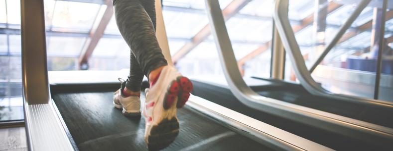 A person’s legs wearing sports trousers and trainers running on a treadmill with windows in the background.