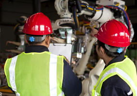 Two workers wearing hi-vis vests and hard hats looking at machinery while checking the contents of a document on a clipboard.