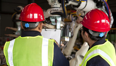 Two workers wearing hi-vis vests and hard hats looking at machinery while checking the contents of a document on a clipboard.