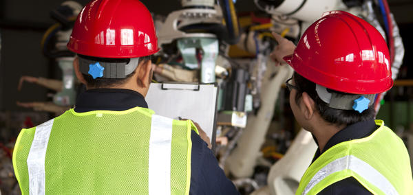 Two workers wearing hi-vis vests and hard hats looking at machinery while checking the contents of a document on a clipboard.