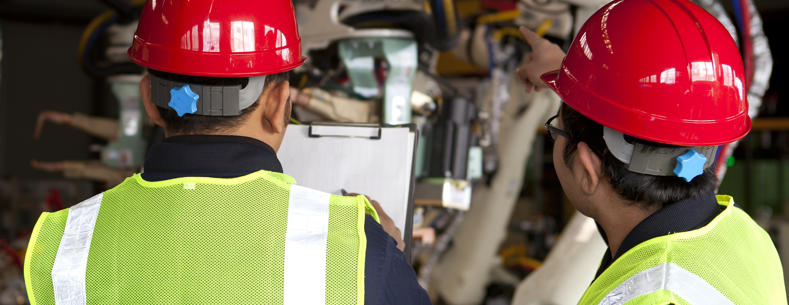 Two workers wearing hi-vis vests and hard hats looking at machinery while checking the contents of a document on a clipboard.