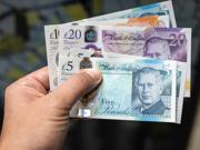 A hand holding a variety of UK bank notes, including £5, £10 and £20.