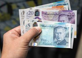 A hand holding a variety of UK bank notes, including £5, £10 and £20.