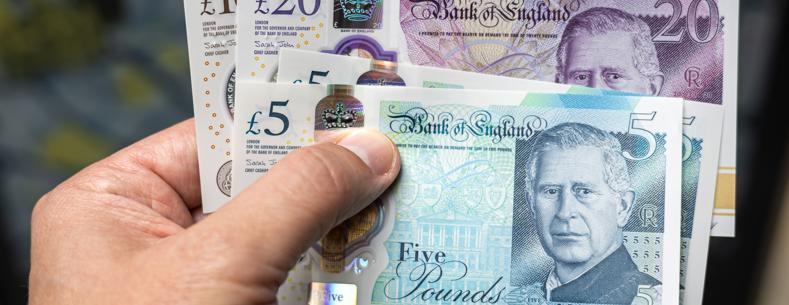 A hand holding a variety of UK bank notes, including £5, £10 and £20.