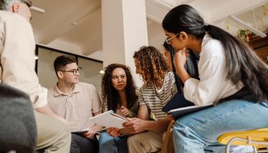 A group of people sitting in a circle in a brightly lit room, looking at papers and talking together during a collaborative discussion.