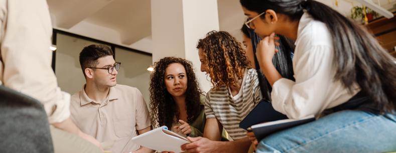 A group of people sitting in a circle in a brightly lit room, looking at papers and talking together during a collaborative discussion.
