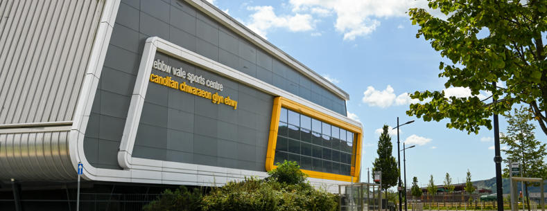 An image of a brand new leisure facility in Ebbw Vale. The leisure centre looks modern and takes the form of a rhomboid, predominantly in grey with glass in a similar shape on the right hand side set in a yellow frame.