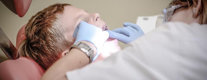 A child sitting in a dentist chair with their mouth open. A dentist wearing glasses and gloves is looking into their mouth.