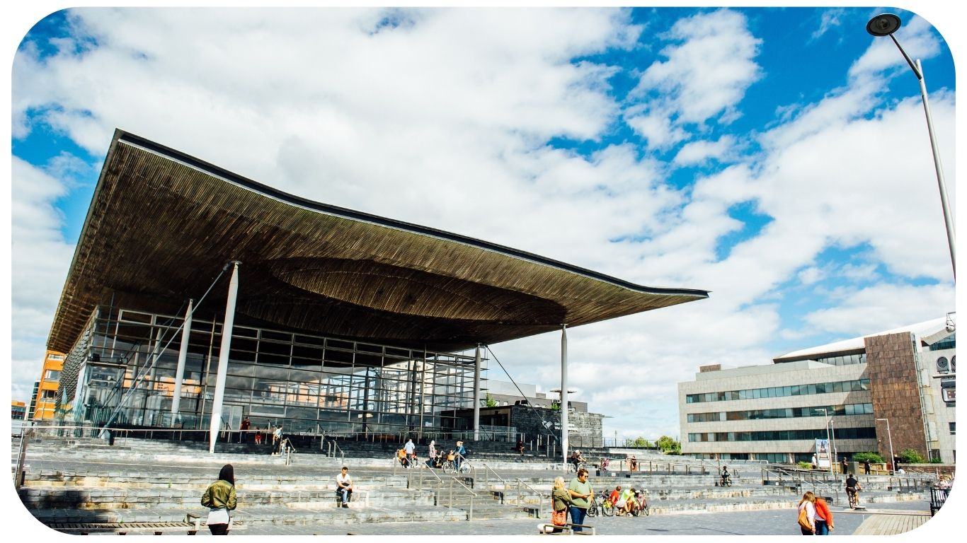 A photograph of the front of the Senedd building in Cardiff Bay. The building is comprised of grey bricks, and some white beams are visible. There is a sign that reads ‘Welsh Parliament’ in the back right hand side. In the foreground, there are pedestrians passing by. Some people are sitting on the steps outside the Senedd building.