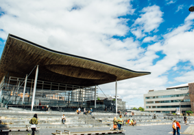 A photograph of the front of the Senedd building in Cardiff Bay. The building is comprised of grey bricks, and some white beams are visible. There is a sign that reads ‘Welsh Parliament’ in the back right hand side. In the foreground, there are pedestrians passing by. Some people are sitting on the steps outside the Senedd building.