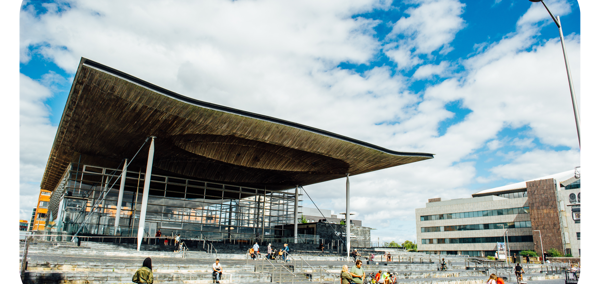 A photograph of the front of the Senedd building in Cardiff Bay. The building is comprised of grey bricks, and some white beams are visible. There is a sign that reads ‘Welsh Parliament’ in the back right hand side. In the foreground, there are pedestrians passing by. Some people are sitting on the steps outside the Senedd building.