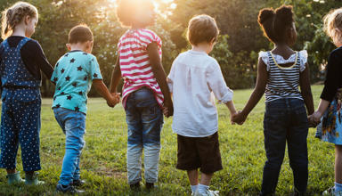 Children playing outdoors on a grassy area, with several children running and interacting in a park-like setting under natural daylight.
