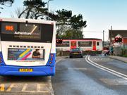 A car waiting at a level crossing while a Transport for Wales train passes. In the foreground a bus is waiting at a bus stop.