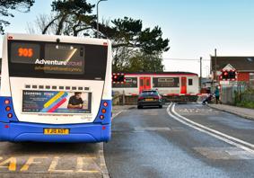 A car waiting at a level crossing while a Transport for Wales train passes. In the foreground a bus is waiting at a bus stop.