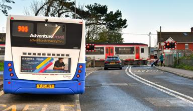 A car waiting at a level crossing while a Transport for Wales train passes. In the foreground a bus is waiting at a bus stop.
