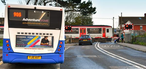 A car waiting at a level crossing while a Transport for Wales train passes. In the foreground a bus is waiting at a bus stop.