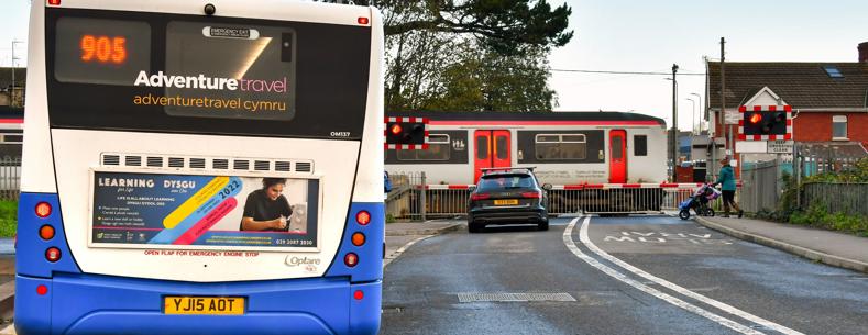 A car waiting at a level crossing while a Transport for Wales train passes. In the foreground a bus is waiting at a bus stop.