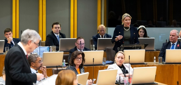 Laura Ann Jones MS stands to address First Minister Mark Drakeford, also standing, during a plenary meeting. Other Members are seated around them.