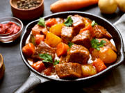 A picture of a warm meat and vegetable stew served in a small pan surrounded by ingredients. 