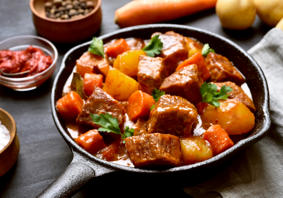 A picture of a warm meat and vegetable stew served in a small pan surrounded by ingredients. 