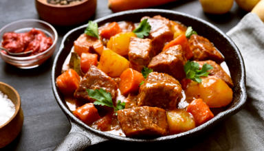 A picture of a warm meat and vegetable stew served in a small pan surrounded by ingredients. 