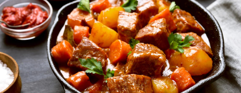 A picture of a warm meat and vegetable stew served in a small pan surrounded by ingredients. 