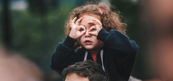 Girl sitting on an adult’s shoulders and using her hands to make binocular shape 