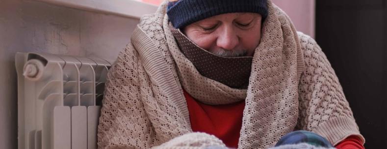 A man huddled next to a radiator wearing a blanket, hat and gloves.