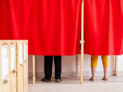 Two people voting in a booth, their legs are visible below red curtains