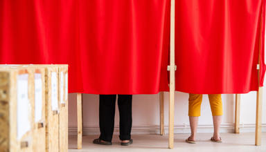 Two people voting in a booth, their legs are visible below red curtains