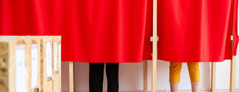 Two people voting in a booth, their legs are visible below red curtains