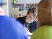 Image of a young woman writing whilst sat at a desk with other people blurred in the background