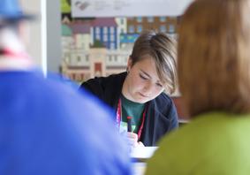 Image of a young woman writing whilst sat at a desk with other people blurred in the background