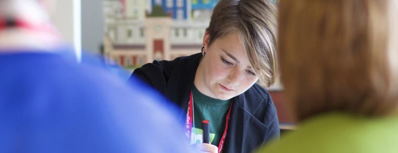 Image of a young woman writing whilst sat at a desk with other people blurred in the background