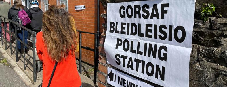 A polling station with a sign saying “polling station” in both English and Welsh clearly visible. A line of people are queuing up to vote.